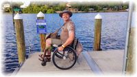 Rollstuhlfahrer steht an einem Fluß. Er schaut in die Kamera und balanciert dabei mit abgehobenen Vorderrädern auf den beiden Hinterrädern.