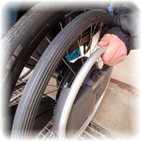 Detailaufnahme eines Rollstuhlhinterrades mit Wheeldrive Antrieb. Die gezeigte Hand am Greifreifen zeigt wie es aussieht, wenn man den Rollstuhl rückwärts fahren möchte.